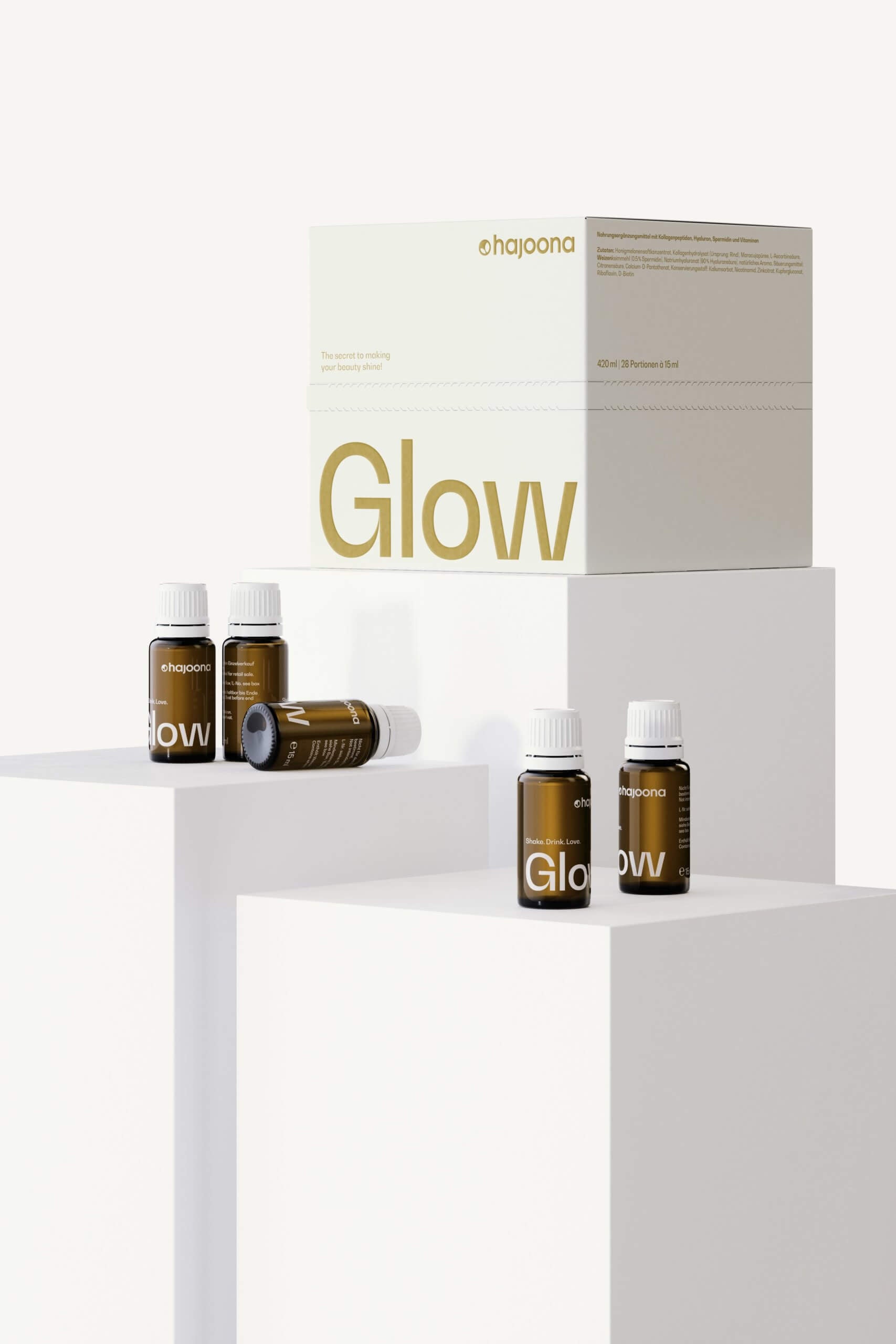 Mehrere kleine braune Glasflaschen mit weißem Deckel stehen auf weißen Podesten. Dahinter eine weiße Verpackung mit goldener Schrift „Glow“, die für Pflege und Schönheit steht. Das Design ist modern und hochwertig.