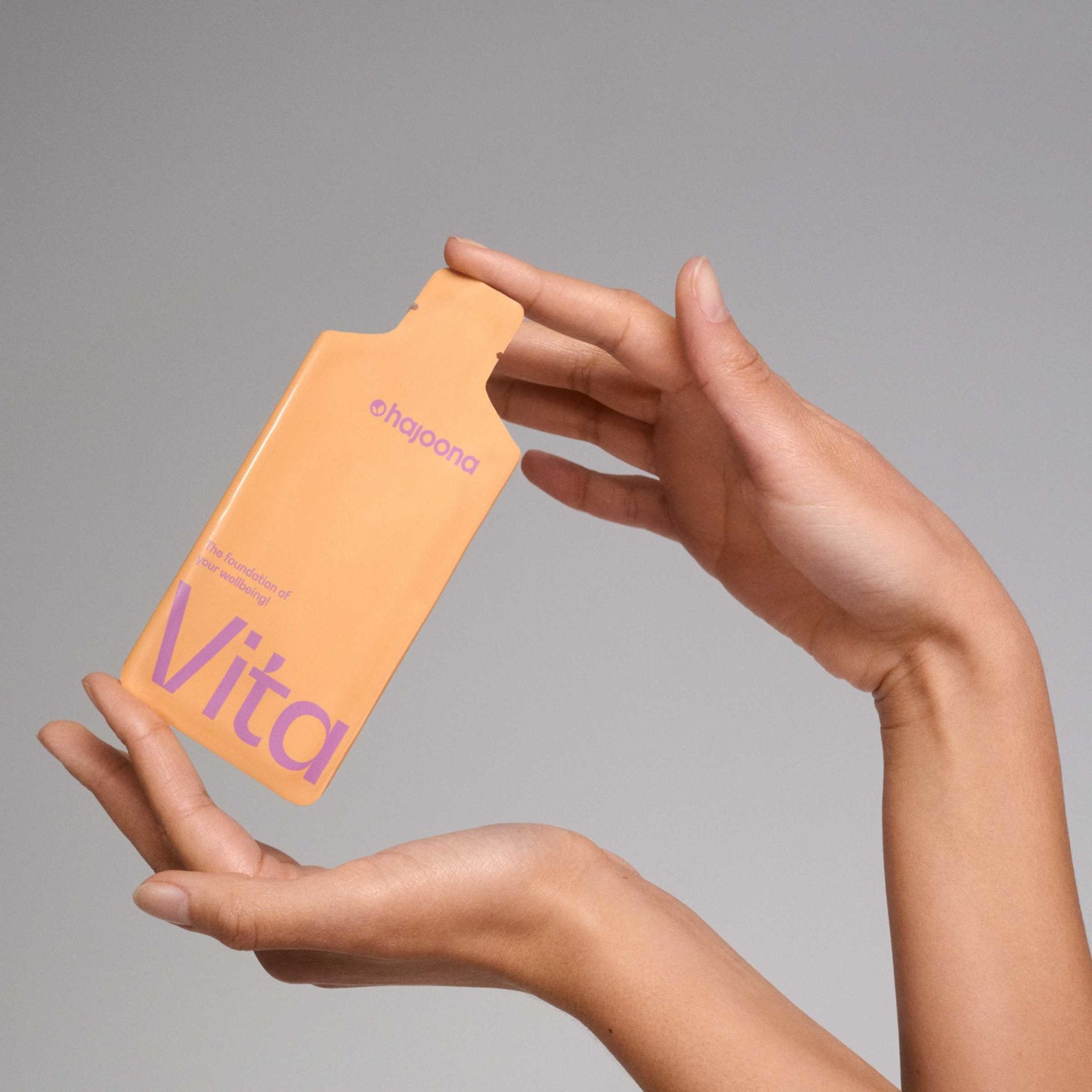 Das Bild zeigt eine Hand, die das Vitality Sachet von hajoona präsentiert. Das Sachet ist in einer Apricot-Farbe gehalten und zeigt den Schriftzug "Vitality" in einem Lila-Ton. Über dem Schriftzug steht "The foundation of your wellbeing!", was auf die wertvolle Unterstützung für das tägliche Wohlbefinden hinweist.