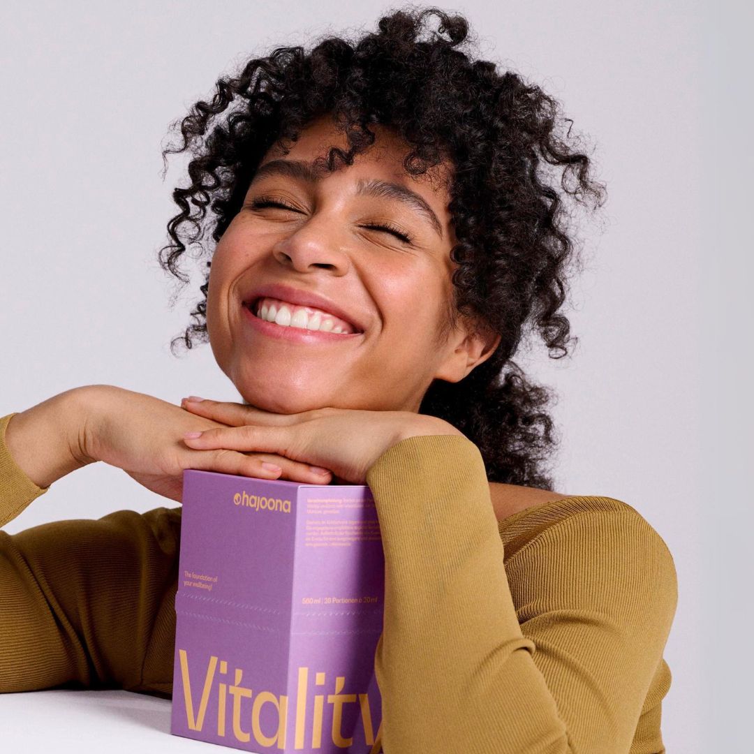 Junge Frau mit lockigem Haar lächelt mit geschlossenen Augen und stützt ihr Kinn auf eine violette hajoona Vitality Packung. Die Szene strahlt Lebensfreude und Wohlbefinden aus.